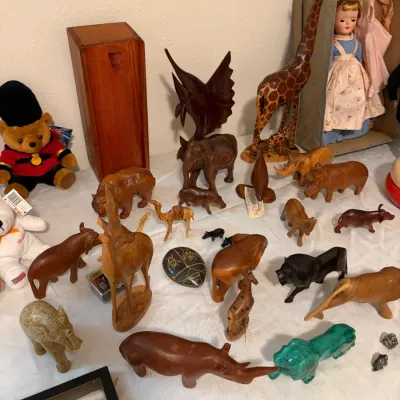 Sale Photo Thumbnail #75: Collection of vintage hand-carved wooden animal figurines, featuring a tall giraffe, elephants, rhinos, and lions, displayed alongside a boxed vintage doll and plush toys.