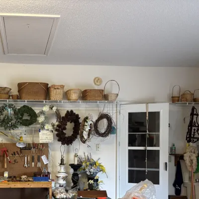 Sale Photo Thumbnail #8: Large assortment of woven wicker baskets and decorative seasonal wreaths displayed on wire shelving above a pegboard workbench with tools and table lamps.