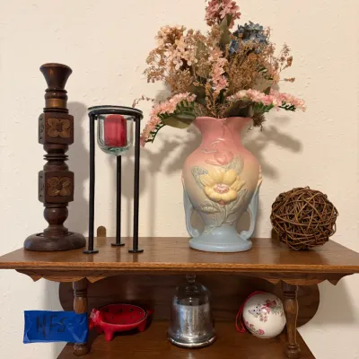 Sale Photo Thumbnail #73: Two-tier oak wall shelf displaying decorative items, featuring a vintage pastel floral ceramic vase with faux flowers, a carved wooden candlestick, and a metal candle holder. The lower shelf holds small collectibles, including a glass cloche and a ceramic