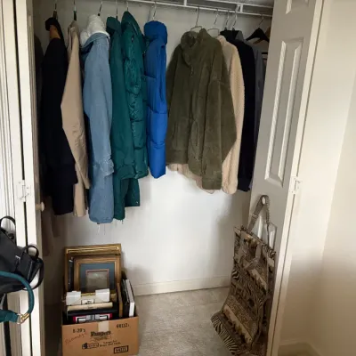 Sale Photo Thumbnail #26: Assorted winter outerwear including fleece pullovers, puffer jackets, and a denim coat hanging in a closet, with a box of framed artwork and handbags nearby.
