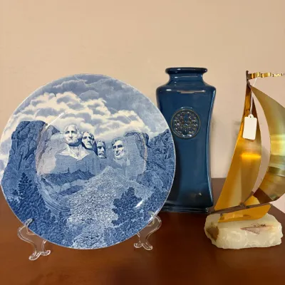 Sale Photo Thumbnail #89: Blue transferware Mount Rushmore souvenir plate on a stand, displayed alongside a blue glazed ceramic vase and a brass sailboat sculpture on a stone base.