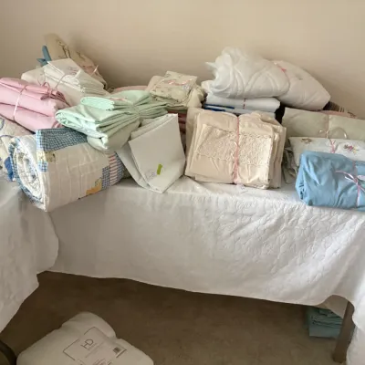 Sale Photo Thumbnail #24: Assorted vintage and contemporary bed linens, including folded sheet sets, quilts, and blankets, neatly arranged on a table.