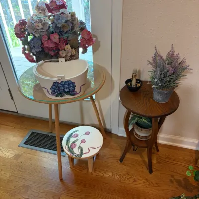 Sale Photo Thumbnail #49: Cottage-style home decor featuring a glass-top tripod side table, a small hand-painted floral stool, and a two-tier wooden accent table decorated with faux floral arrangements.