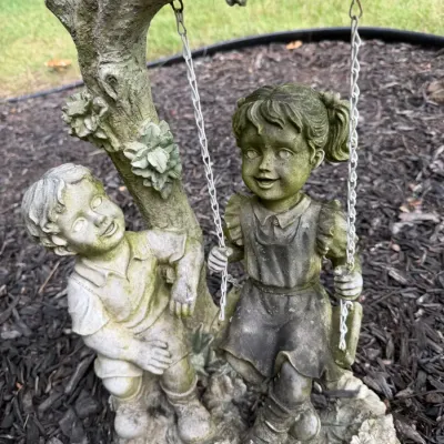 Weathered garden statue of a boy pushing a girl on a swing attached to a tree branch