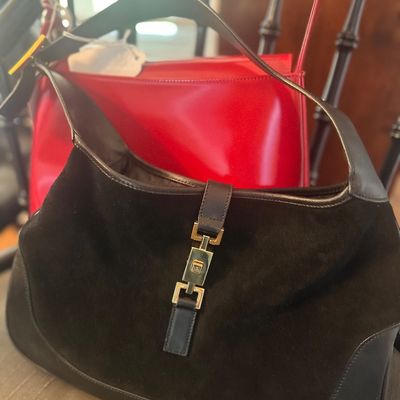 Vintage Gucci Jackie-style shoulder bag in black suede and leather with gold-tone piston closure, shown in front of a red leather handbag.