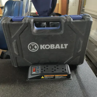 Kobalt hard plastic tool carrying case with blue latches, shown alongside a black power strip featuring three outlets and two USB ports.