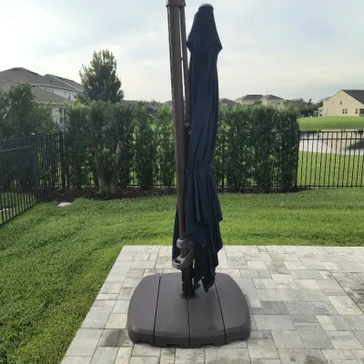 Large cantilever patio umbrella with a dark blue canopy, brown metal frame, and a heavy-duty plastic base, shown closed on a paved patio.