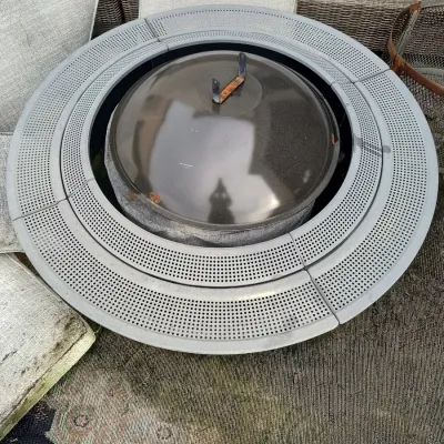 Round metal fire pit with a perforated grey surround and a black domed lid, showing some signs of outdoor wear.