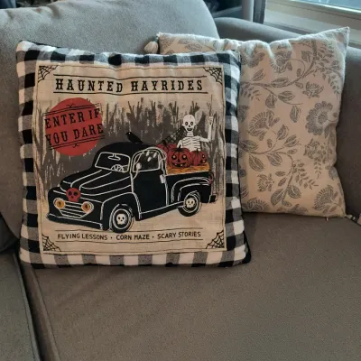 Halloween-themed throw pillow featuring a black and white buffalo plaid border and a vintage-style graphic of a haunted hayride truck,...