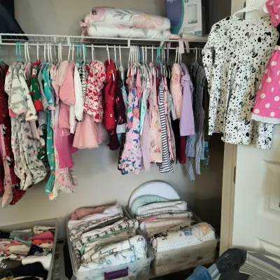 Assorted baby and toddler clothing hanging in a closet, alongside plastic bins filled with folded baby blankets and linens.
