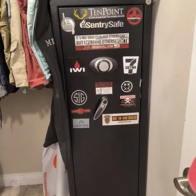 SentrySafe tall black gun safe with a combination lock, lever handle, and various firearm-related stickers on the door.