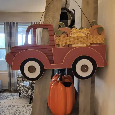 Rustic wooden hanging sign shaped like a red pickup truck carrying pumpkins, displayed alongside a large orange ceramic pumpkin on a...