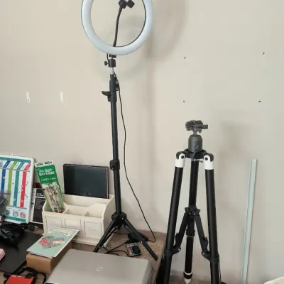 Modern ring light on an adjustable tripod stand alongside a black and white camera tripod, shown with an HP laptop and desk accessories.
