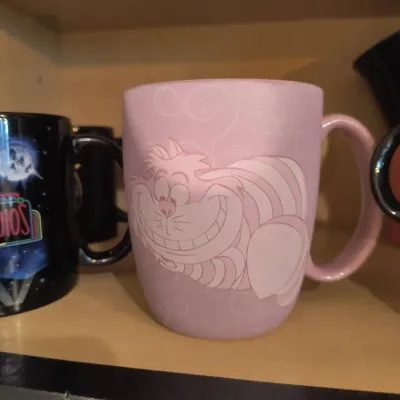 Disney Cheshire Cat ceramic coffee mug in pastel pink, featuring a stylized illustration of the character on a shelf alongside other mugs.
