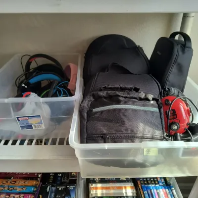 Assorted electronics accessories, including headphones and black carrying cases, organized in clear plastic bins on a shelf above a...