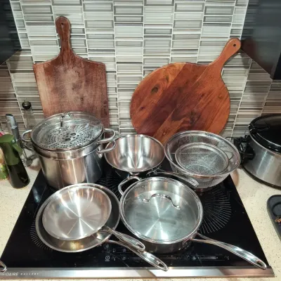 Assorted stainless steel cookware including pots, pans, a steamer insert, and mesh strainers, displayed alongside two wooden cutting...