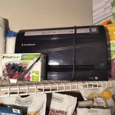 Black FoodSaver vacuum sealer machine shown with a box of heat-seal pre-cut bags on a wire shelf.