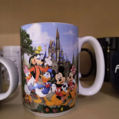 Disney theme park souvenir mug featuring Mickey Mouse, Minnie Mouse, Donald Duck, Daisy Duck, Goofy, and Pluto in front of Cinderella...