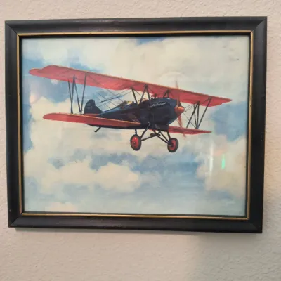 Framed vintage-style print depicting a classic blue and red biplane flying through clouds, housed in a dark wood frame with gold inner trim.