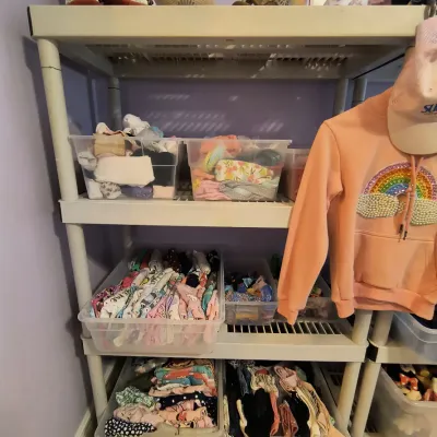 White plastic shelving unit holding clear storage bins filled with children's clothing, alongside a pink rainbow hoodie and various...