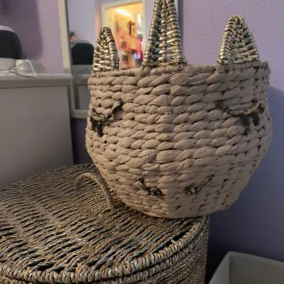 Whimsical woven basket shaped like a cat or unicorn face with metallic gold ears, sitting atop a larger lidded wicker storage hamper.