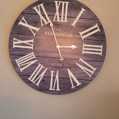 Large round farmhouse-style wall clock featuring a gray faux-wood plank face, white Roman numerals, and the text 'FARMHOUSE EST....