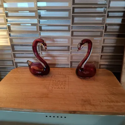 Pair of vintage Murano-style cranberry art glass swan figurines, displayed on a wooden bread box lid.