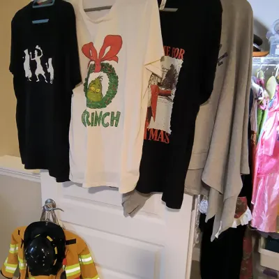 Assorted graphic t-shirts hanging on a door, including a Grinch holiday shirt, alongside a child's firefighter costume with helmet.