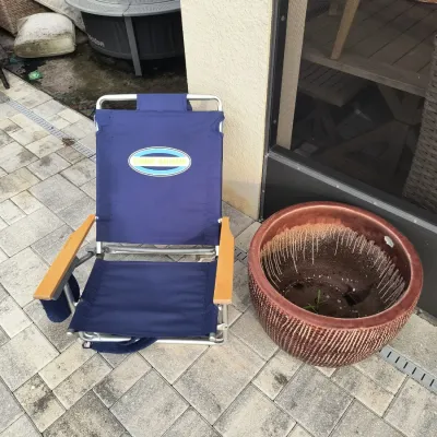 Tommy Bahama folding beach chair in navy blue with wooden armrests, shown next to a large textured ceramic planter on a paved patio.
