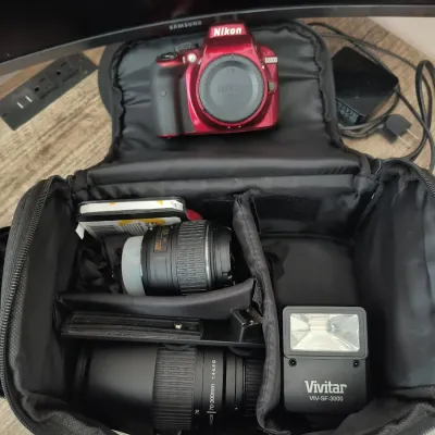 Red Nikon D3300 DSLR camera body shown with a black padded carrying bag containing two Nikkor lenses and a Vivitar VIV-SF-3000 flash unit.