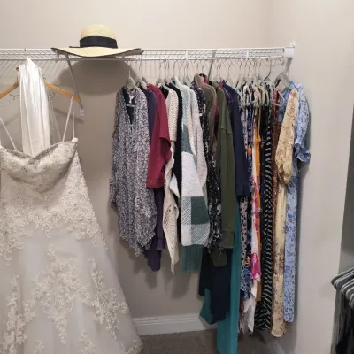 Strapless white lace wedding gown displayed alongside a closet rack of women's clothing, including dresses, sweaters, and blouses.