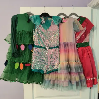 Four children's dress-up costumes hanging on a door, including a green Christmas tree dress, a sequined teal and pink romper, a...