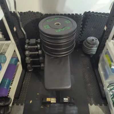CAP Barbell flat weight bench stacked with 25 lb bumper plates, shown alongside a dumbbell rack and other home gym equipment.