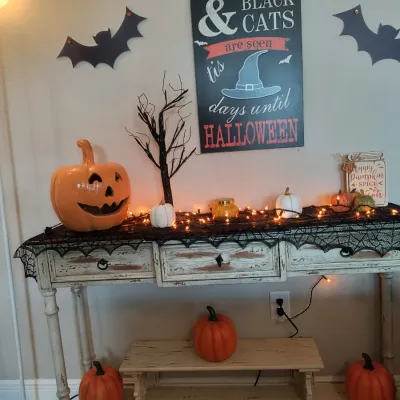 Distressed white wooden console table decorated with Halloween items, including a large ceramic jack-o'-lantern, faux pumpkins, a...