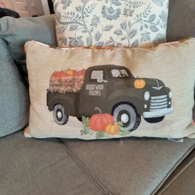 Two decorative throw pillows on a gray sofa, featuring a rectangular pillow with an embroidered vintage green truck and pumpkin design,...