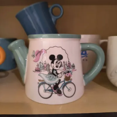 Disney Minnie Mouse ceramic watering can-shaped mug featuring Minnie riding a bicycle, displayed on a shelf with other mugs.