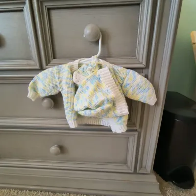 Hand-knit baby sweater and matching hat set in a pastel yellow, blue, and white variegated yarn, displayed on a hanger.