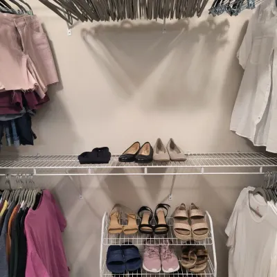 Assorted women's shoes, including sandals, flats, and sneakers, displayed on a white wire rack and shelf alongside hanging clothing.