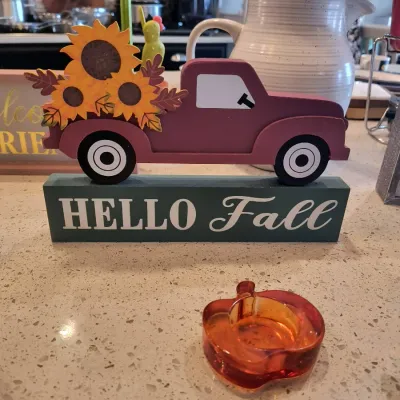 Wooden "Hello Fall" tabletop sign featuring a red pickup truck filled with sunflowers, paired with a small orange glass...