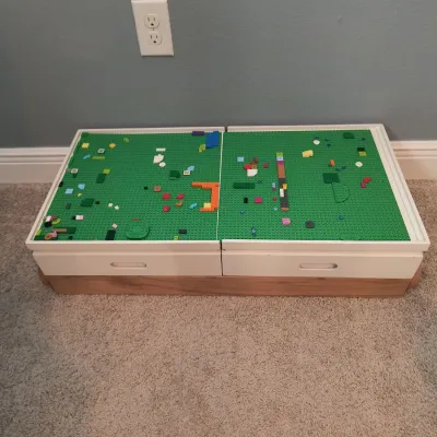 Low-profile wooden toy storage chest featuring two white pull-out drawers and a top surface fitted with green Lego baseplates.