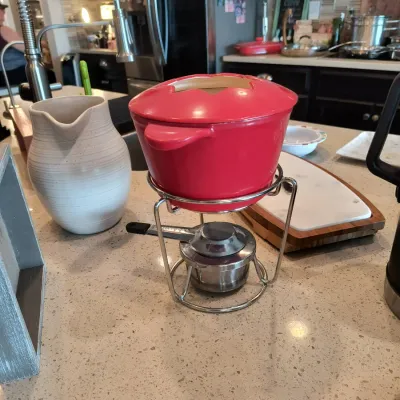 Red ceramic fondue pot with a matching lid, sitting on a wire stand with a metal burner, shown alongside a white ceramic pitcher on a...