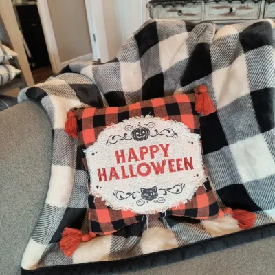 Halloween-themed decorative throw pillow with "Happy Halloween" text, orange and black buffalo plaid pattern, and corner...