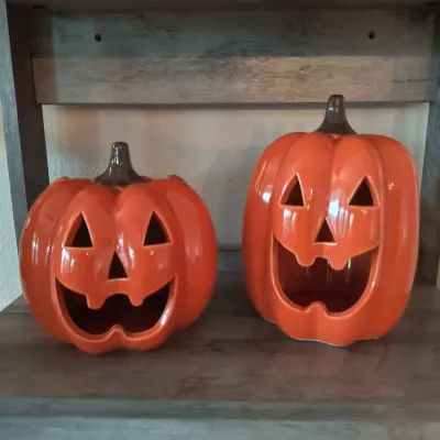 Pair of glossy orange ceramic jack-o'-lantern decorations in two different sizes, featuring carved-out faces and brown stems.