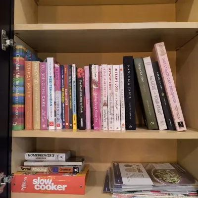 Collection of modern cookbooks and recipe binders, including titles like "Magnolia Table" and "Wheat Belly," arranged...