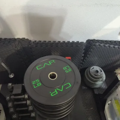 CAP Barbell 25 lb bumper plates stacked on a rack, alongside smaller weight plates, dumbbells, and interlocking foam gym floor mats.