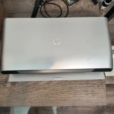 HP silver laptop shown closed, resting on a wooden surface next to a tripod and remote control.