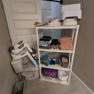 White plastic four-tier shelving unit holding towels, linens, and children's bath accessories, shown alongside a toddler potty...
