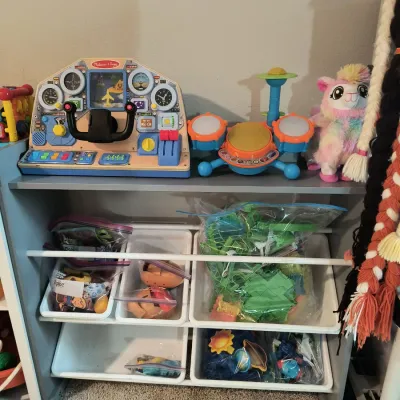 Children's toy storage organizer shelf filled with various toys, including a Melissa & Doug dashboard activity center, a toy...