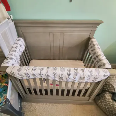 Modern wooden baby crib in a weathered gray finish, featuring a high paneled backboard and patterned rail guards.
