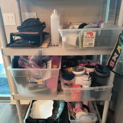 White plastic utility shelving unit holding various kitchen items, including plastic storage bins, travel mugs, a cast iron press, and...